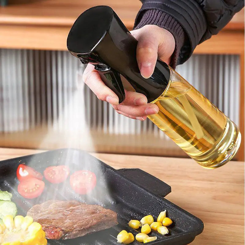 Hand holding a black Oil & Vinegar Spray Bottle, misting oil over a sizzling steak and vegetables on a grill pan. Ideal for precise cooking and grilling.